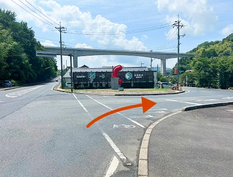 玉造温泉街への入口を示す交差点の風景。中央には「玉造温泉」と書かれた建物があり、赤いオブジェが立っている。右手に進むよう、オレンジ色の矢印が画像に重ねて描かれている。背景には高架道路が見える。