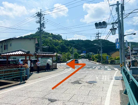 玉造温泉街の中心部へと続く交差点の写真。橋を渡り、オレンジ色の矢印に従って左折するよう示されている。交差点の左手には休憩所のような建物があり、人々が立っているのが見える。