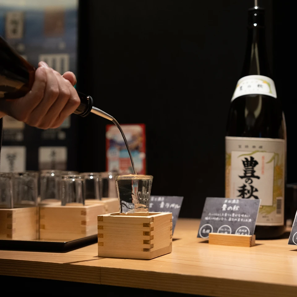 旅館のバーカウンターで、升に入ったグラスにお酒が注がれている画像です。銘柄が書かれたお酒の瓶や、飲み比べができるように複数の升とグラスが並べられており、夕食時に様々な種類のお酒を楽しめる雰囲気が伝わってきます。