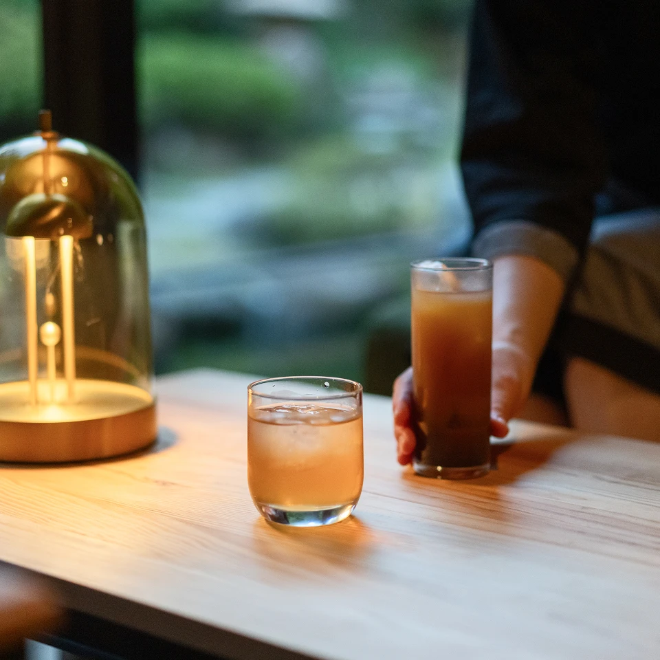 バーラウンジの木製テーブルに、飲み物が2つ並べられている画像です。おしゃれな照明が灯る落ち着いた雰囲気の中で、飲み物を片手にゆったりと過ごす様子が伝わってきます。