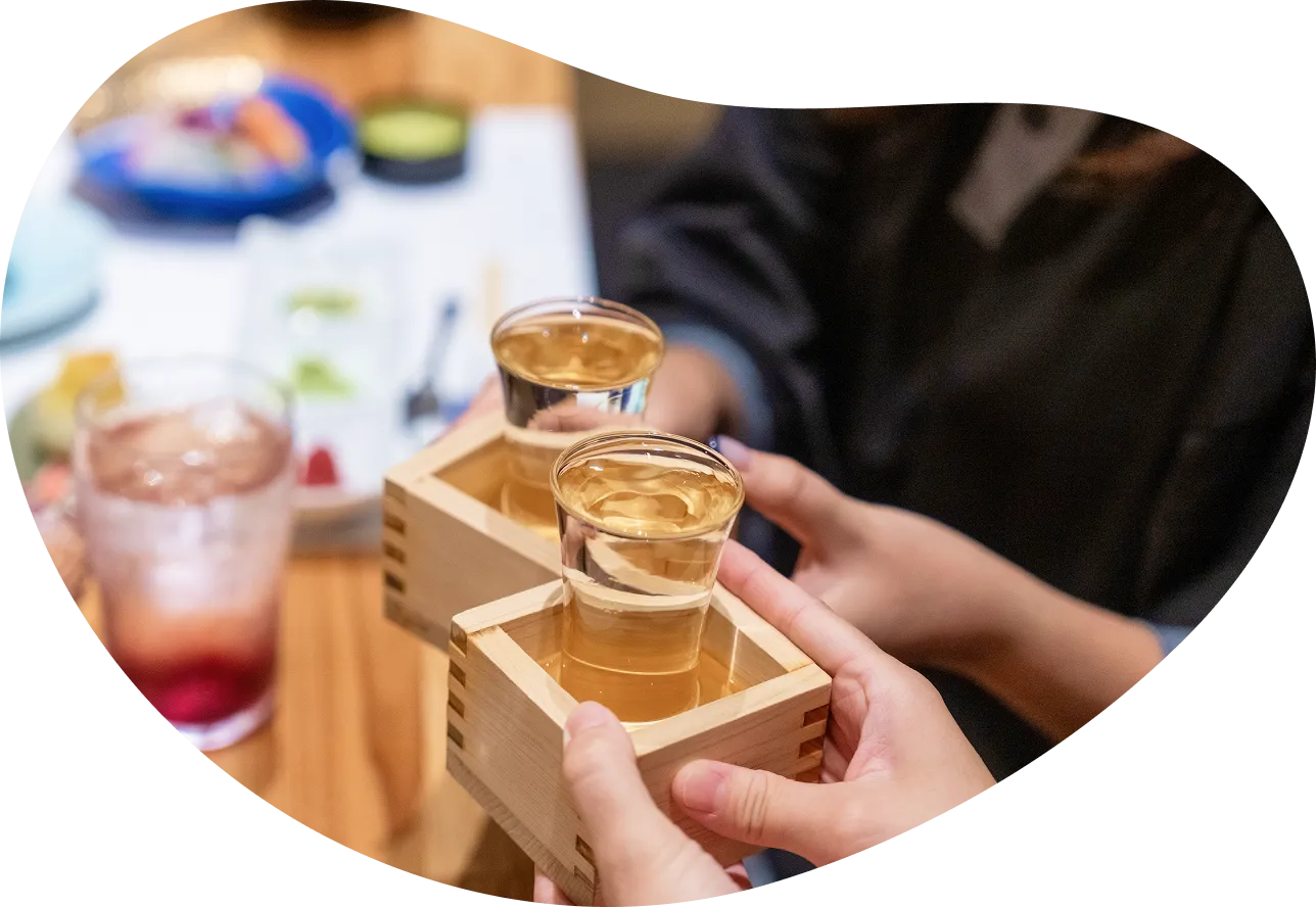 旅館の食事処で、お酒が入ったグラスと升を持った手が乾杯しようとしている画像です。楽しい食事の時間が始まる様子が伝わってきます。