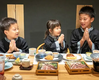 旅館の食事処で、3人の子どもたちが館内着を着て食事をしている画像です。皆が手を合わせて「いただきます」のポーズをしており、食事を心待ちにしている楽しそうな雰囲気が伝わってきます。