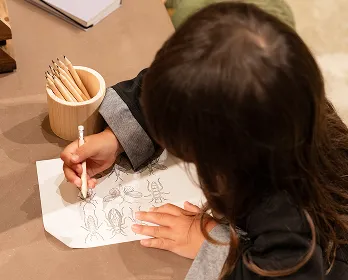 キッズスペースで、女の子が鉛筆を手に絵を描いている画像です。昆虫らしきイラストが描かれた紙に熱中している様子が伝わってきます。