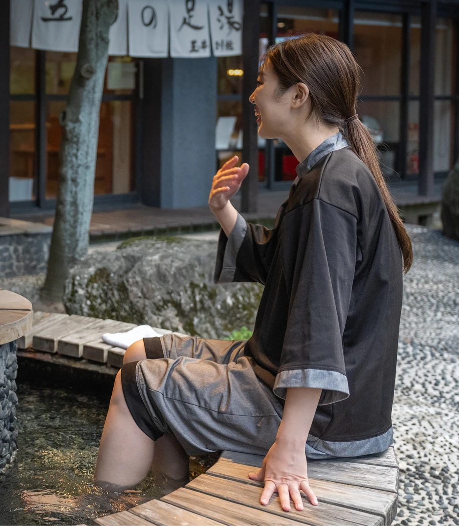 屋外にある足湯で、館内着を着た女性2人が笑顔で談笑している画像です。足湯の湯気に癒されながら、リラックスして過ごす様子が伝わってきます。