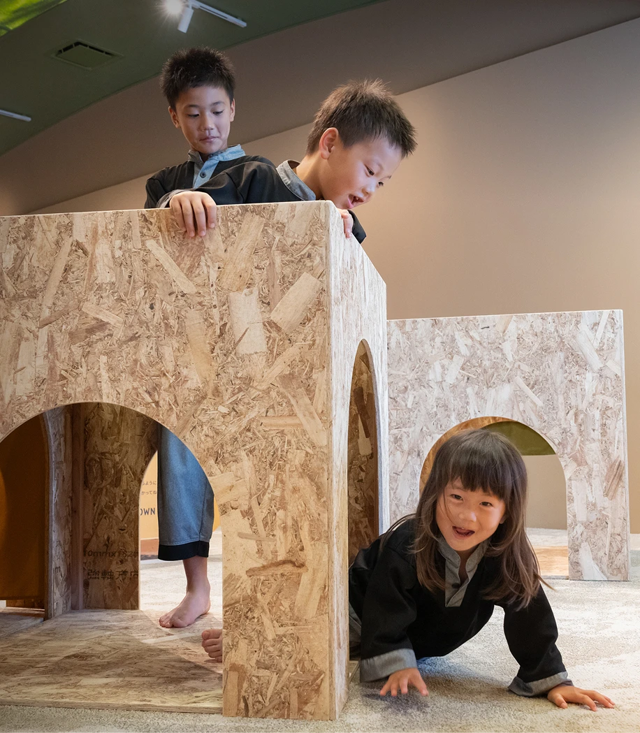 合板で作られたトンネルのような遊具で遊ぶ子どもたちの画像です。下から顔を出して笑う女の子と、その上から顔をのぞかせる男の子が写っており、楽しそうな笑い声が聞こえてくるようです。